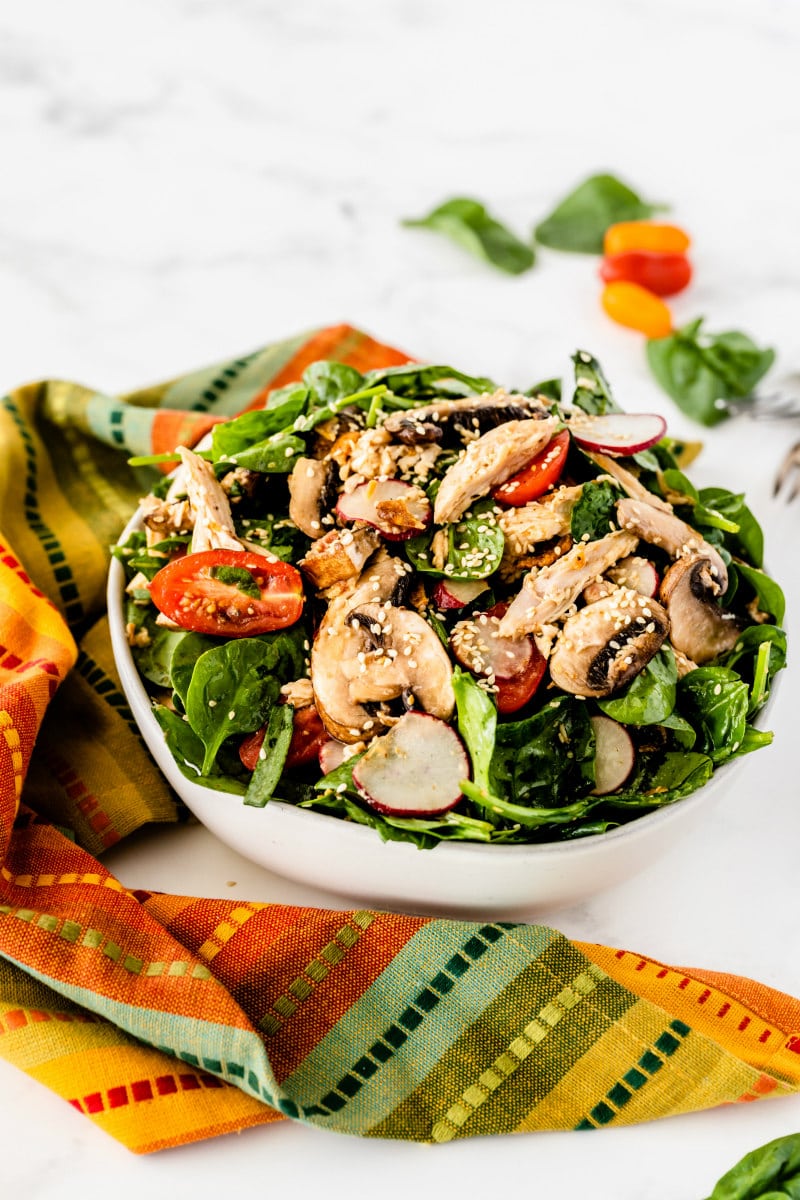 mandarin chicken spinach salad in a white bowl with a bright and colorful patterned cloth napkin displayed in front