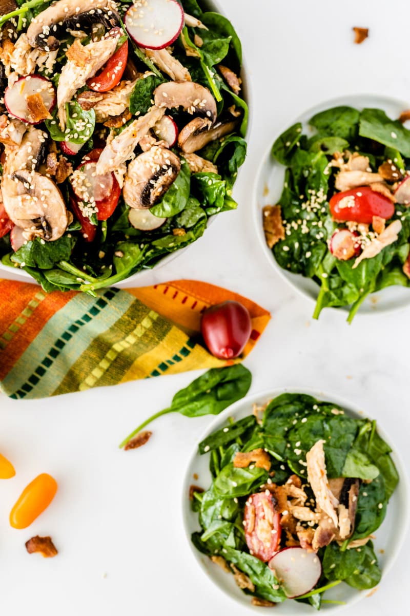 overhead shot of mandarin chicken spinach salad in a bowl with two servings of the salad on plates. Colorful cloth napkin displayed too.