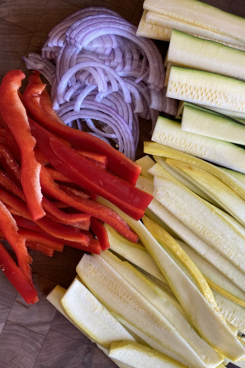 sliced vegetables displayed on a cutting board: red bell peppers, red onion, yellow squash and zucchini