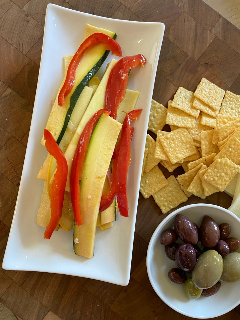 serving sweet and sour summer vegetables on a white plate displayed on a cutting board with crackers and a white bowl of olives