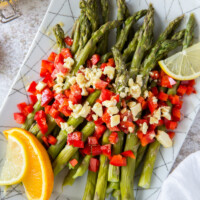a white plate set on a marble background with chilled asparagus that are topped with feta vinaigrette with chopped red bell pepper and garnished with lemons and oranges