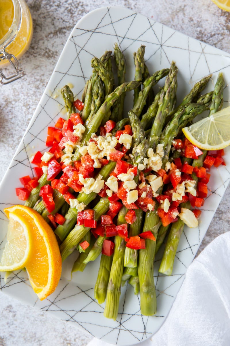 a white plate set on a marble background with chilled asparagus that are topped with feta vinaigrette with chopped red bell pepper and garnished with lemons and oranges