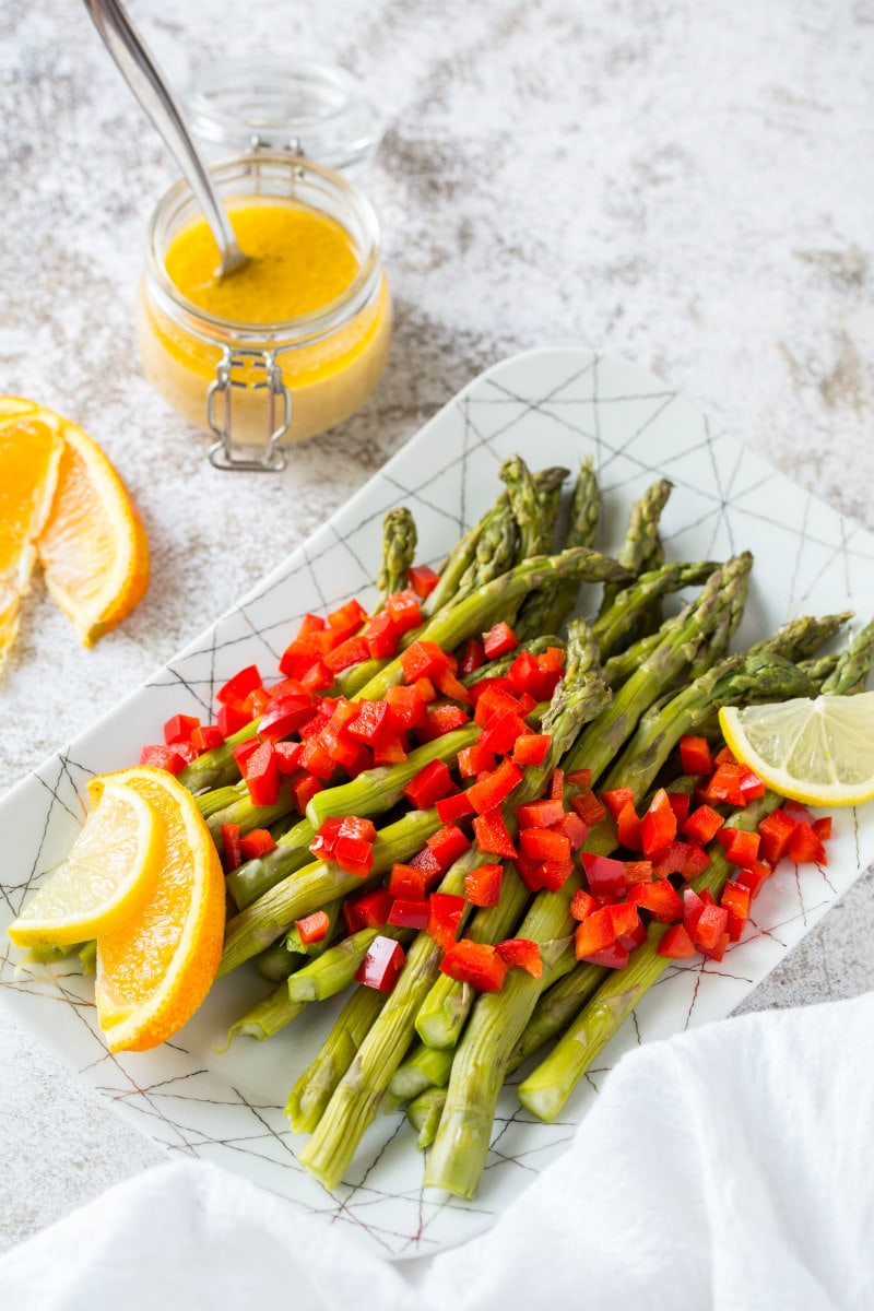 a white plate set on a marble background with chilled asparagus that are topped with feta vinaigrette with chopped red bell pepper and garnished with lemons and oranges. glass of vinaigrette on the side