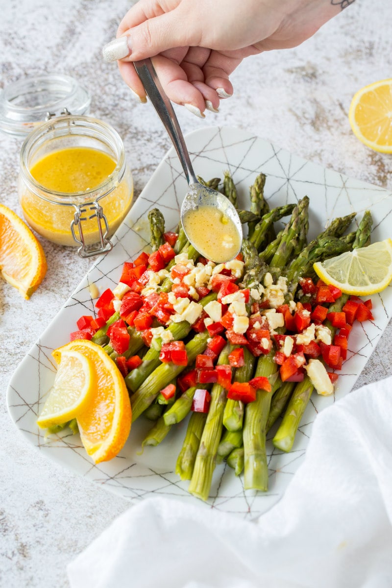 a white plate set on a marble background with chilled asparagus showing a hand spooning feta vinaigrette on top with chopped red bell pepper and garnished with lemons and oranges