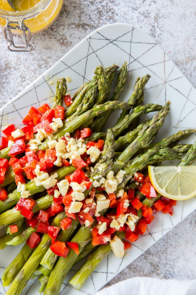 a white plate set on a marble background with chilled asparagus that are topped with feta vinaigrette with chopped red bell pepper and garnished with lemons and oranges