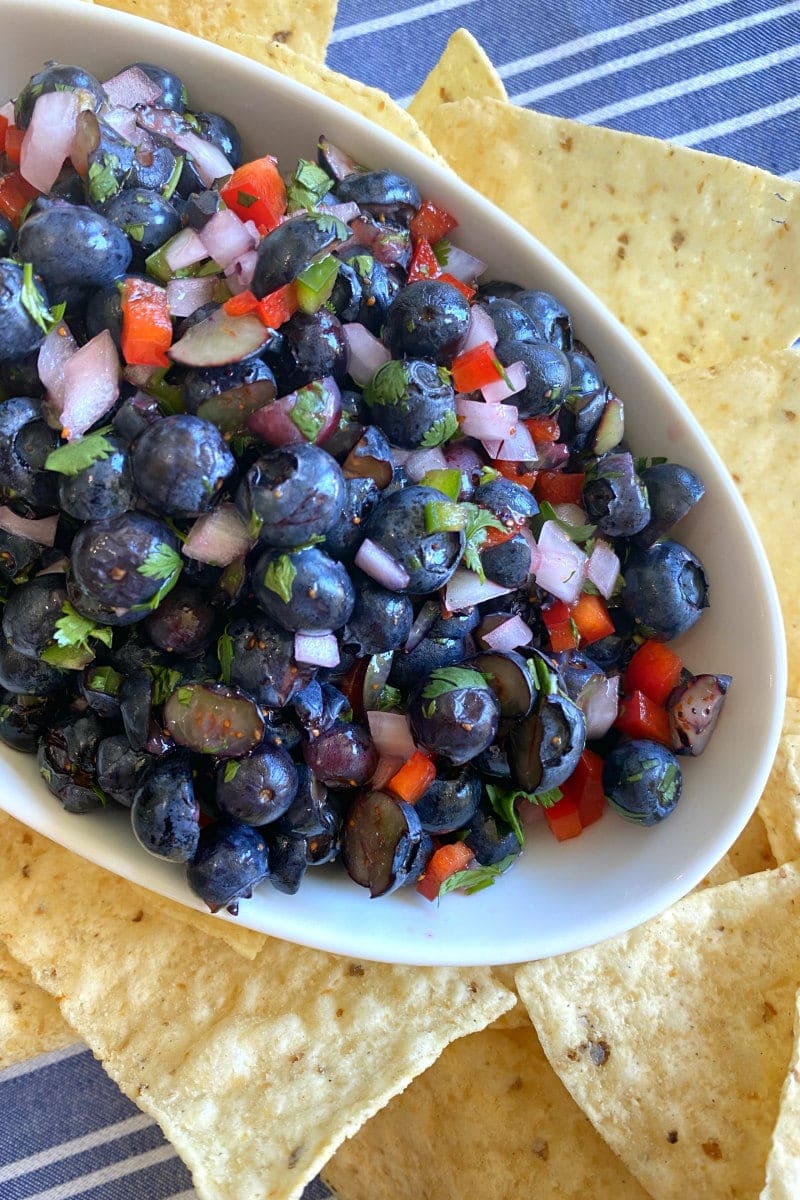 blueberry salsa in a white bowl surrounded by tortilla chips on a white/blue striped napkin