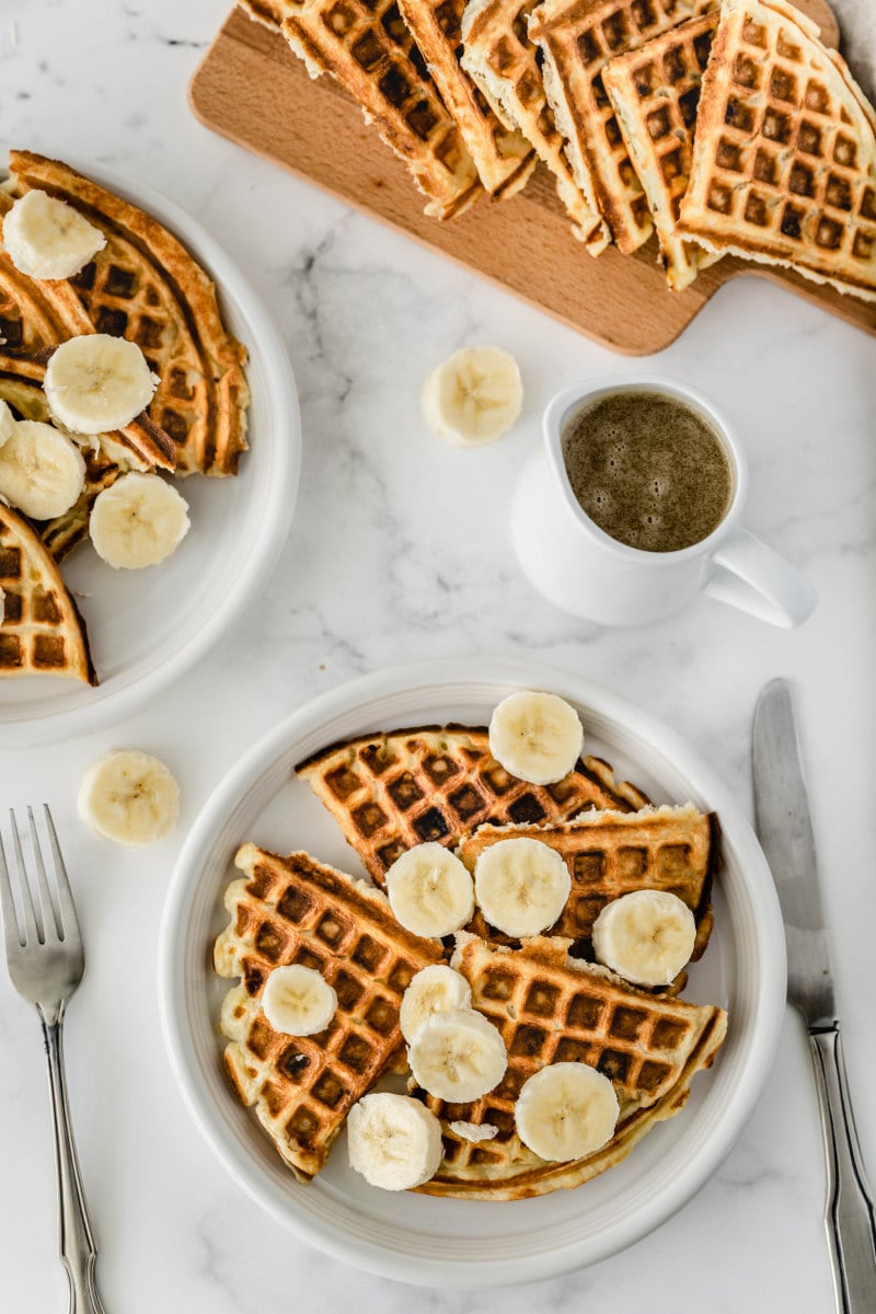 white plates with banana waffles on top with sliced bananas and a pitcher of syrup