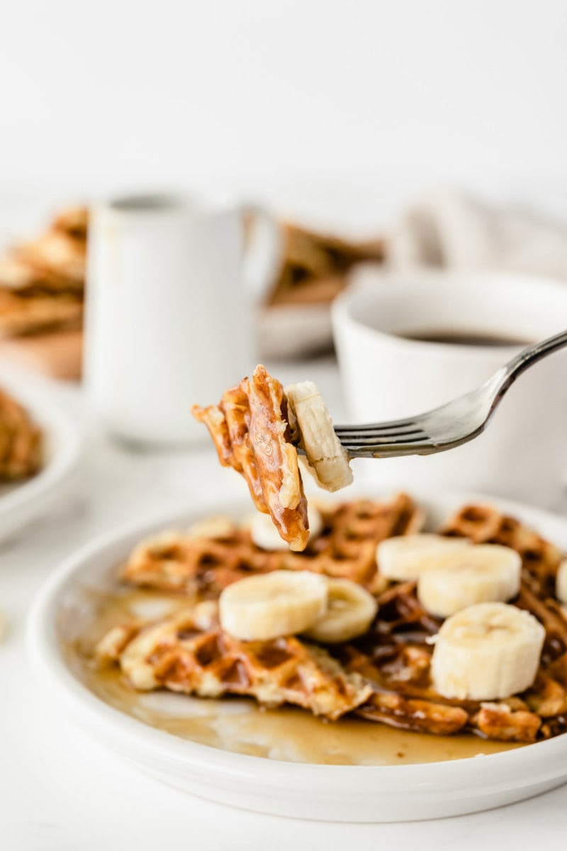 forkful of waffle above a plate of banana waffles. Cups of coffee in the background