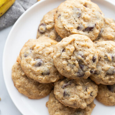 overhead shot of banana walnut cookies stacked on a white plate with a peek of a banana in the background