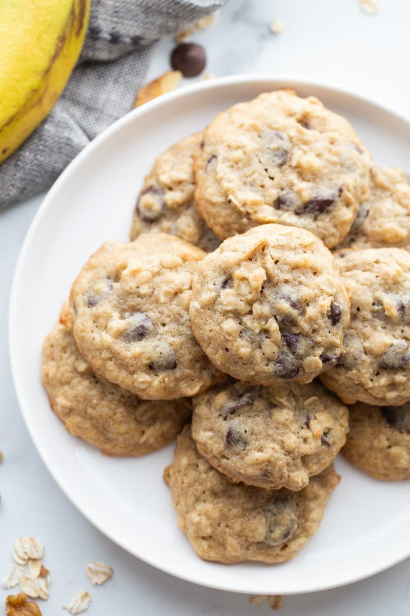 overhead shot of banana walnut cookies stacked on a white plate with a peek of a banana in the background