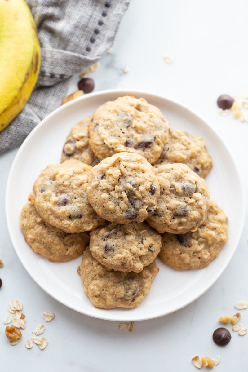 overhead shot of banana walnut cookies stacked on a white plate with a peek of a banana in the background