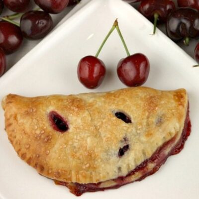 fresh cherry turnover on a white plate with a couple of fresh cherries on the plate. fresh cherries in the background
