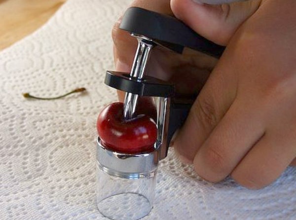 showing process of using a cherry pitter to get the pit out of a cherry- fingers holding cherry pitter and pressing down on cherry