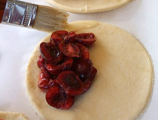 showing process of making cherry turnovers with cherry filling on a pastry and using a brush to brush edges with water