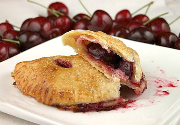 two cherry turnovers on a white plate with one cut open to see the inside. Fresh cherries in the background