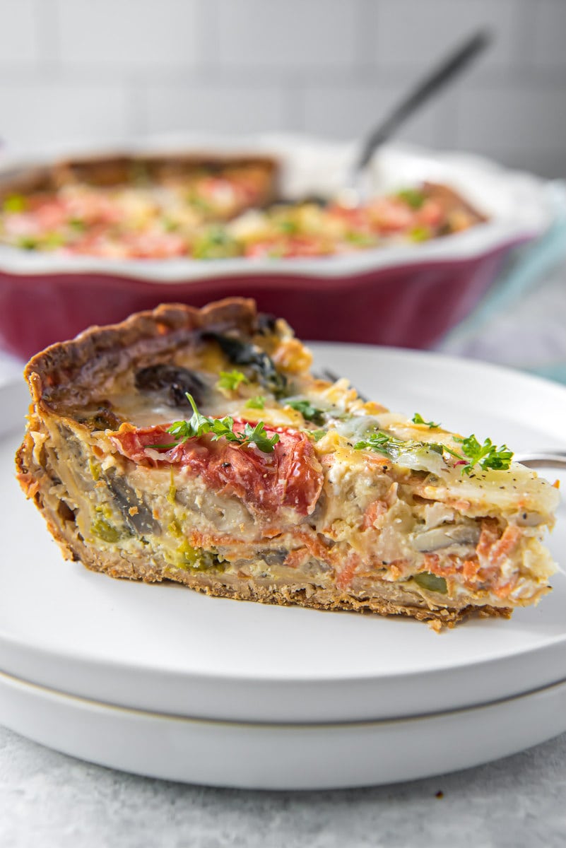 slice of garden vegetable quiche on a white plate with full quiche in a red pie dish in the background