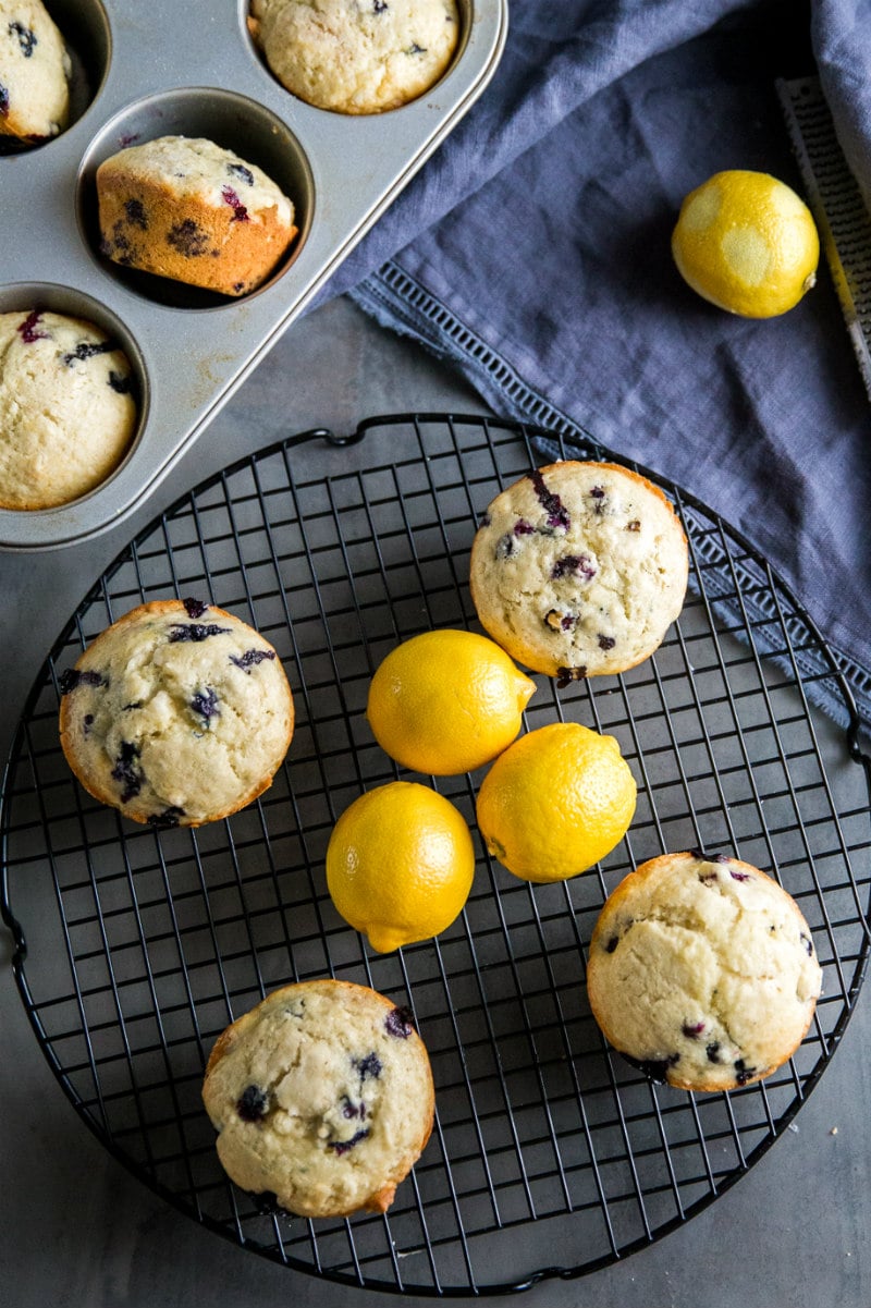 overhead shot of lemon blueberry muffins on a cooling rack with fresh lemons. muffins in a muffin pan on the side, and a blue cloth napkin displayed.