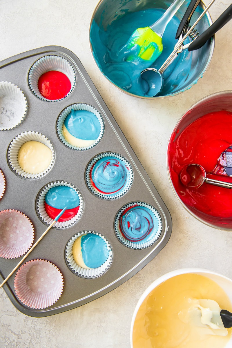 red white and blue cupcake batter in a cupcake pan with bowls of each color batter along the sides. A long wooden stick used to do the swirling.
