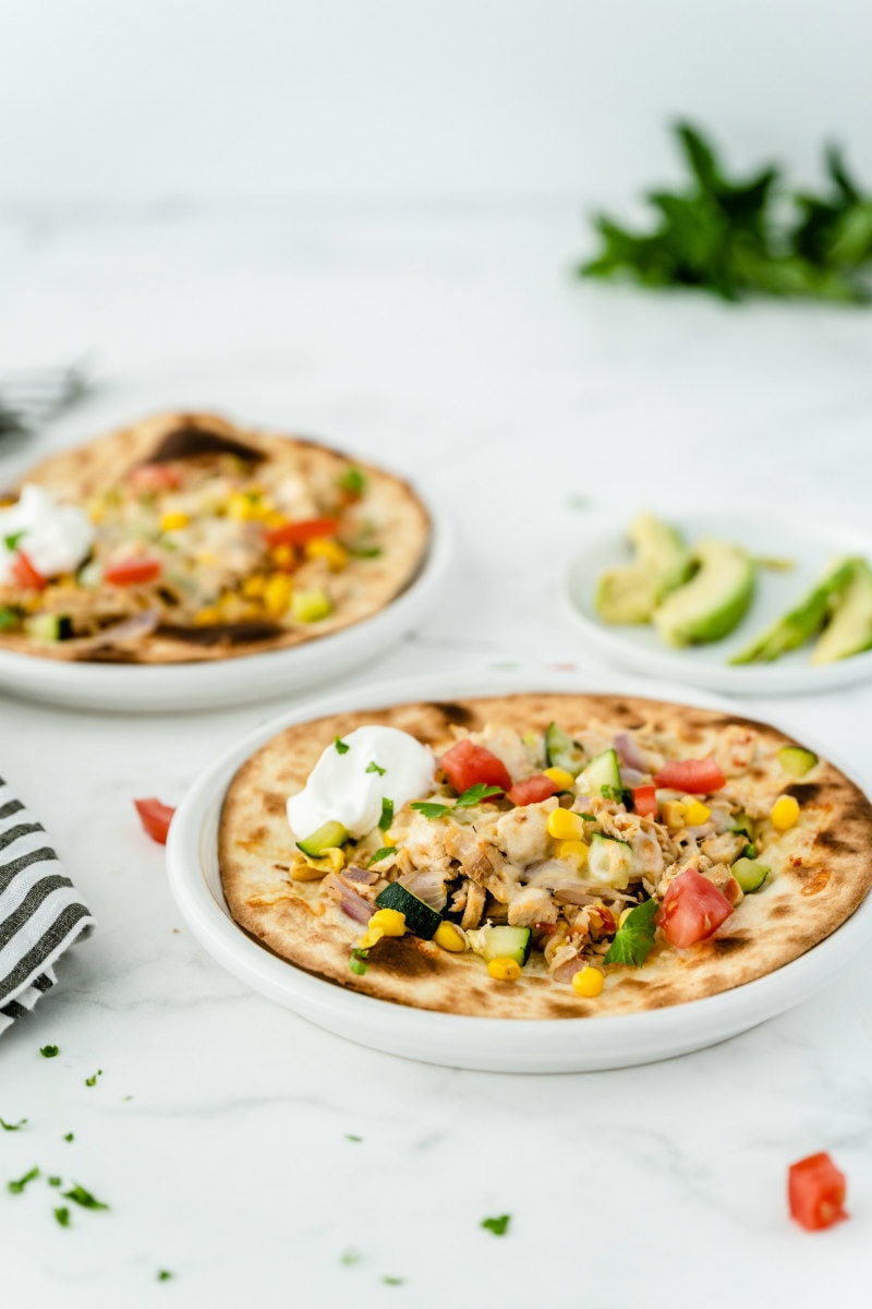 two white plates with chicken and summer vegetable tostadas with a small white plate of sliced avocado in the background and a blue/white striped napkin peeking in on the side