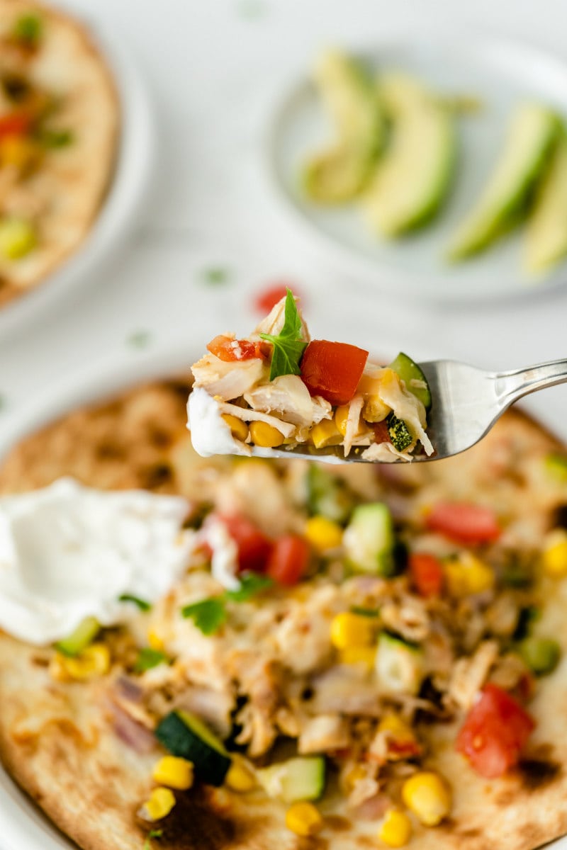 fork full of chicken and summer vegetable tostada above the tostada. peek at another tostada in the background and a small white plate of sliced avocado