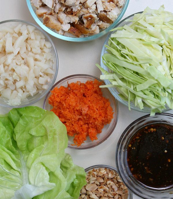 overhead shot of Ingredients for Sweet and Spicy Chicken Lettuce Wraps in glass bowls- chicken, water chestnuts, carrots, sliced cabbage, sauce, peanuts and lettuce leaves