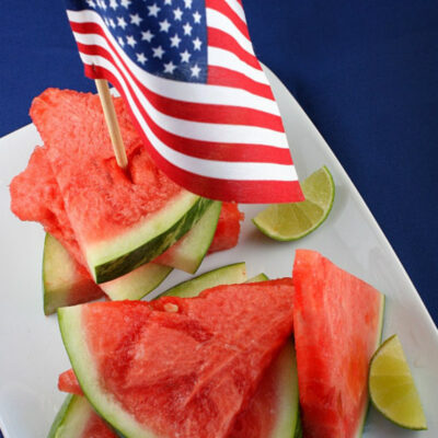 tequila soaked watermelon wedges on a white platter with lime wedges and an american flag stuck into the top