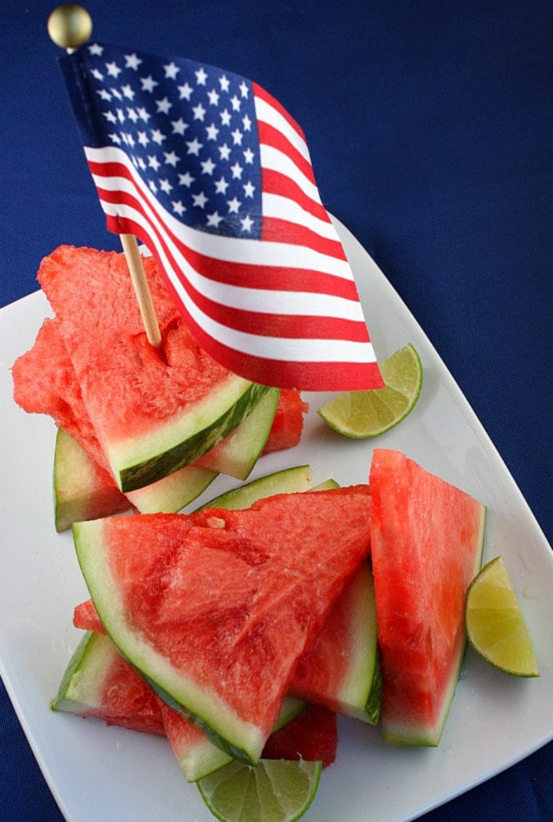 tequila soaked watermelon wedges on a white platter with lime wedges and an american flag stuck into the top