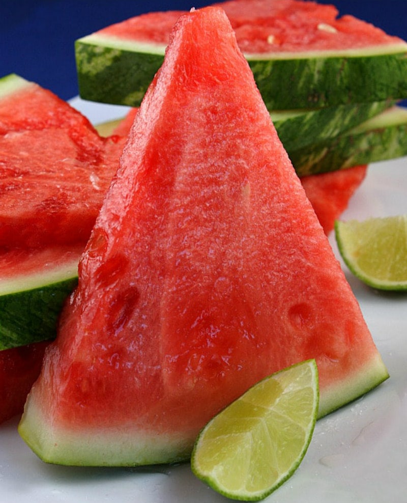 tequila soaked watermelon wedge standing up on its side with a lime wedge in front. more watermelon in background