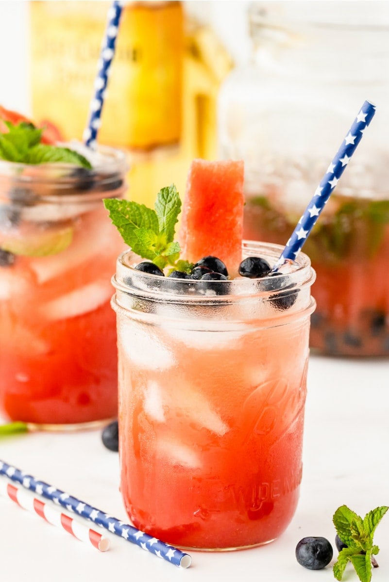 watermelon tequila cocktails in mason jars garnished with fresh blueberries and mint. striped straws. bottle of tequila in the background