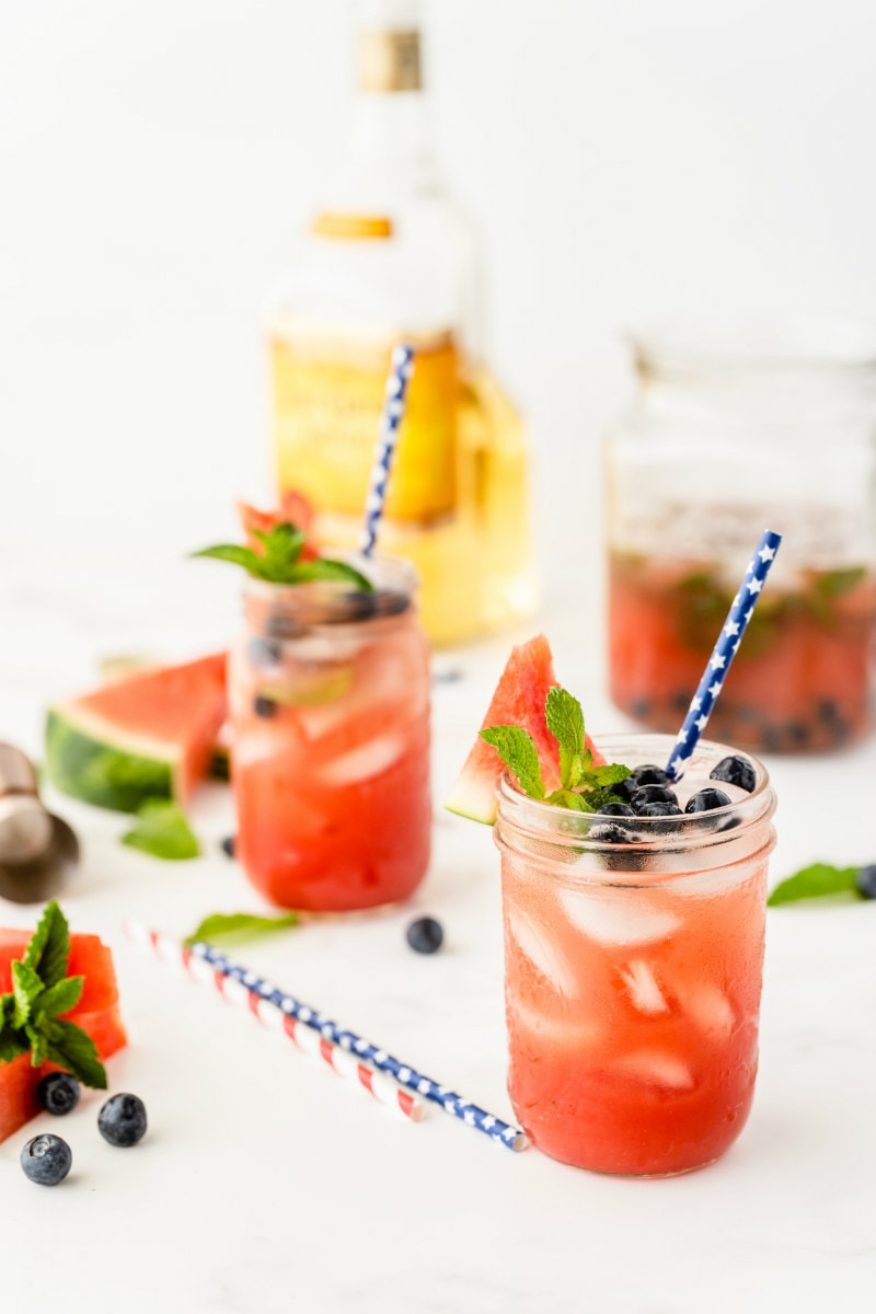 watermelon tequila cocktails in mason jars garnished with fresh blueberries and mint. striped straws. bottle of tequila in the background