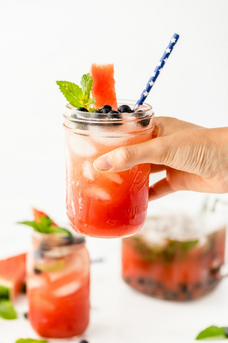 hand holding a watermelon tequila cocktail in the air above more of the same cocktails