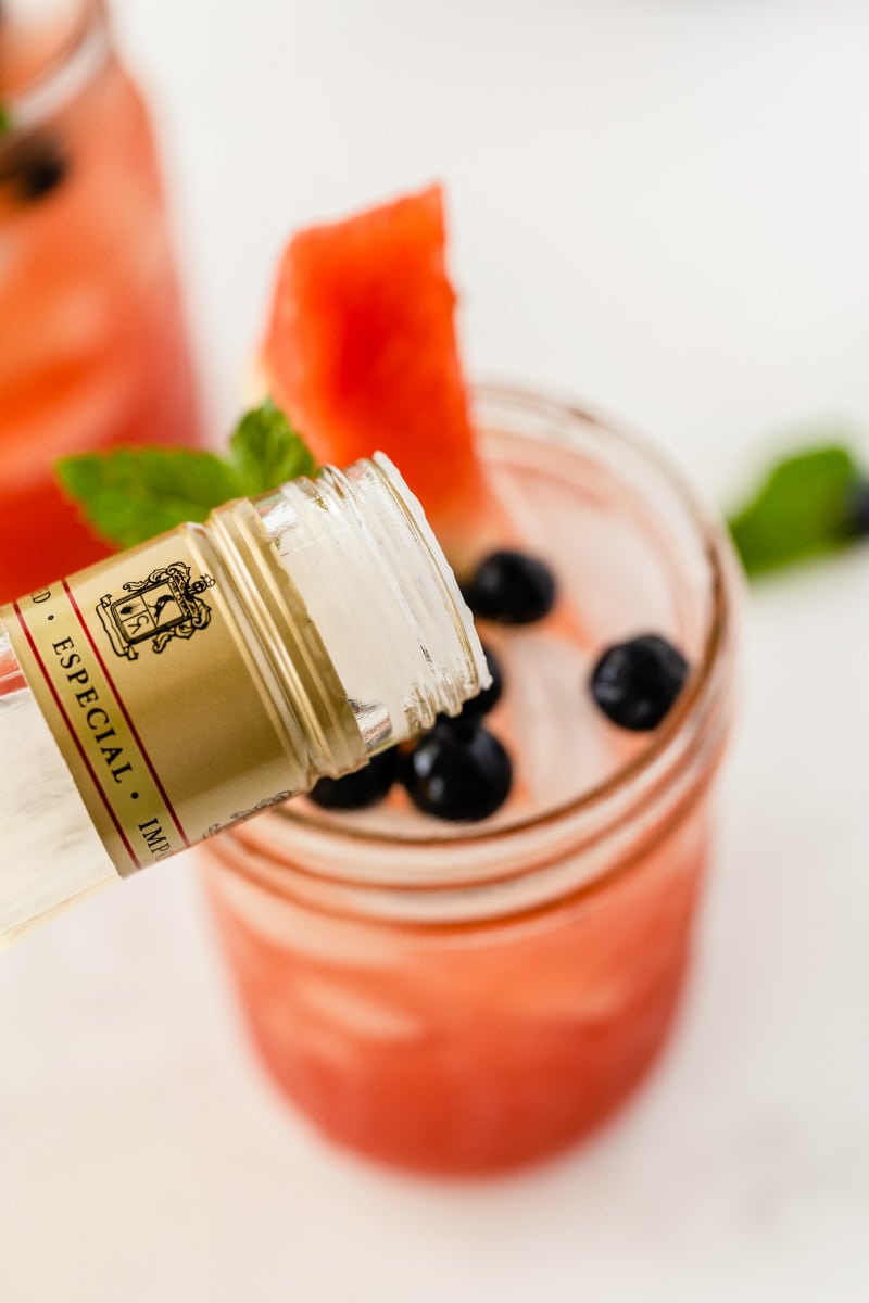 pouring tequila into watermelon tequila cocktail in a mason jar. garnished with fresh blueberries and watermelon wedge.