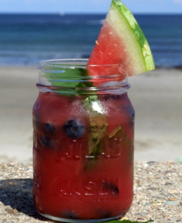 watermelon tequila cocktail in a mason jar with watermelon wedge garnish. beach view in the background