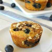 Blueberry Cinnamon Muffin on a white plate with more muffins on a platter in the background with a white and blue striped napkin