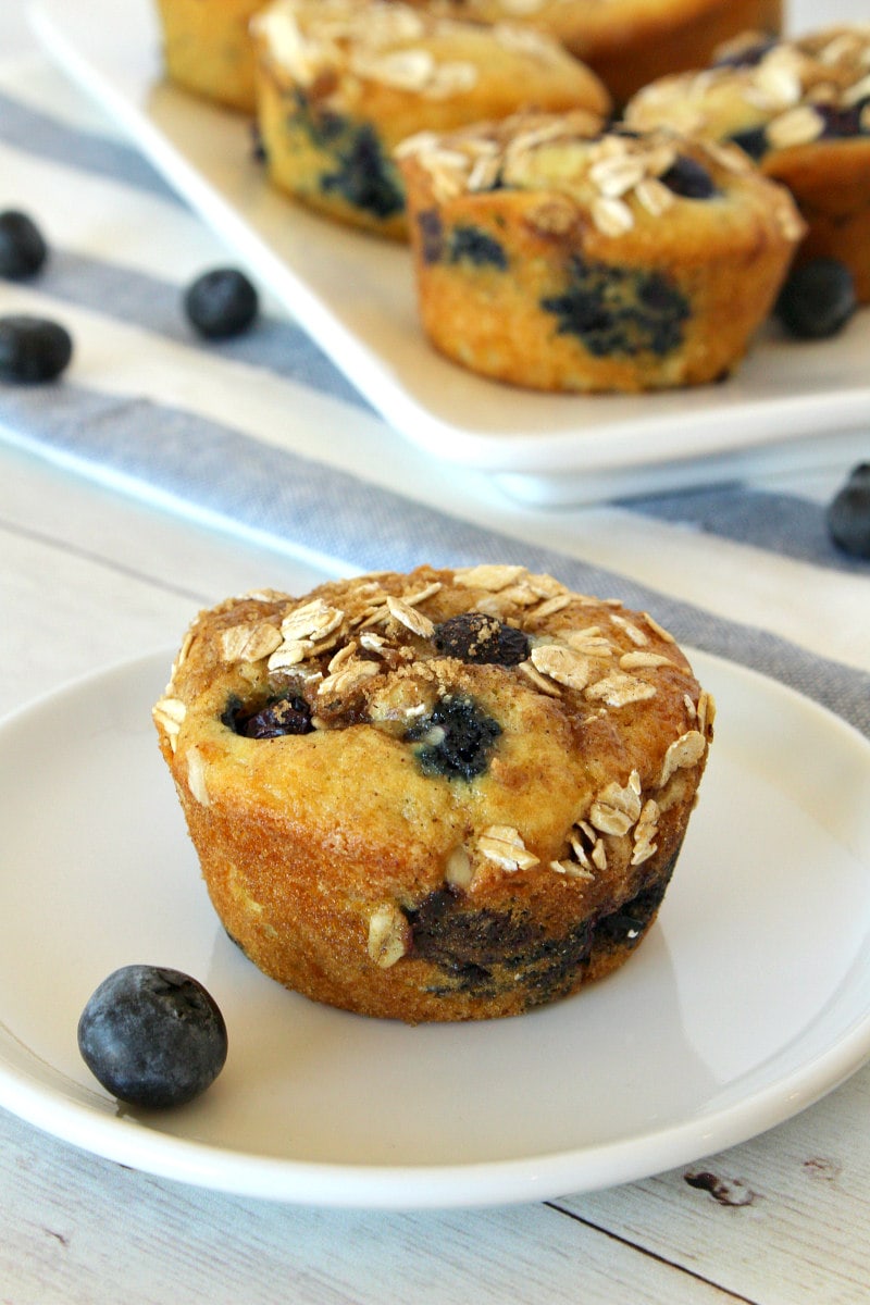 Blueberry Cinnamon Muffin on a white plate with more muffins on a platter in the background with a white and blue striped napkin