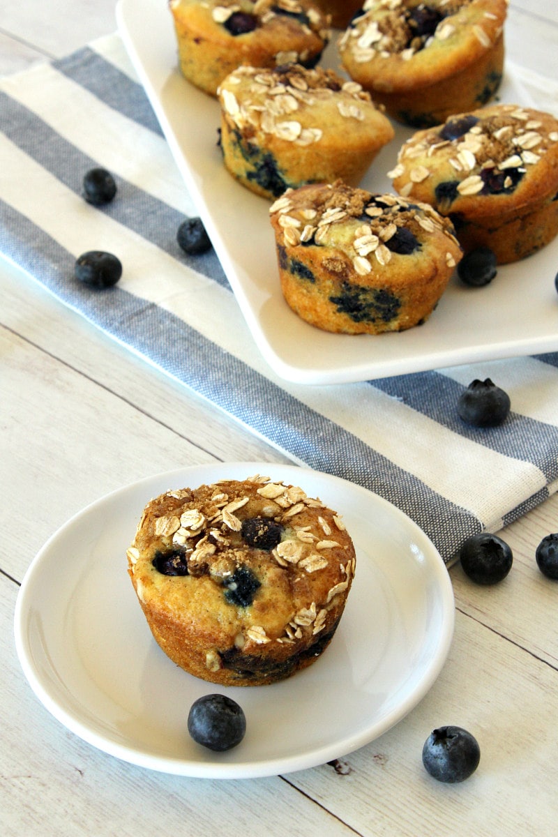 serving Blueberry Cinnamon Muffins on a white plate with more muffins on a white platter in the background with a white and blue striped napkin