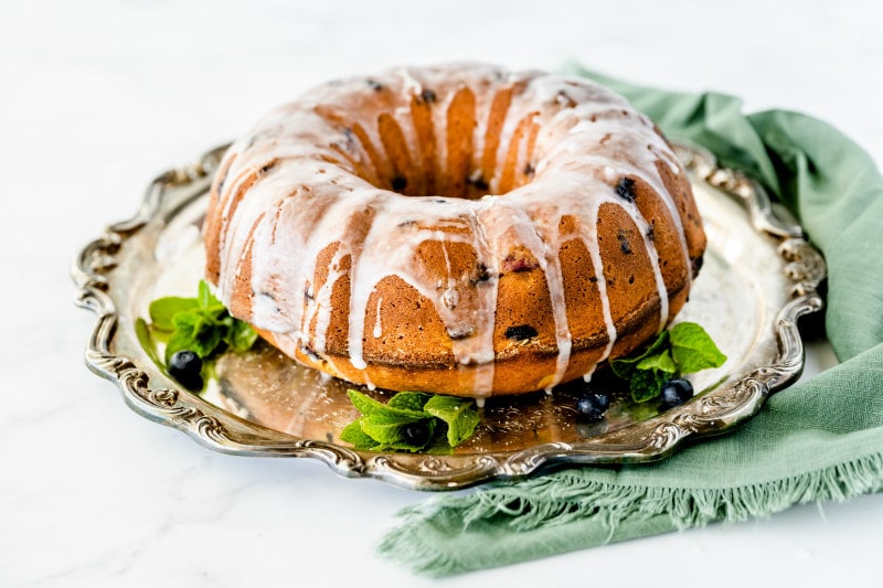 blueberry pound cake on a silver platter with glaze on top, garnished by mint leaves and fresh blueberries