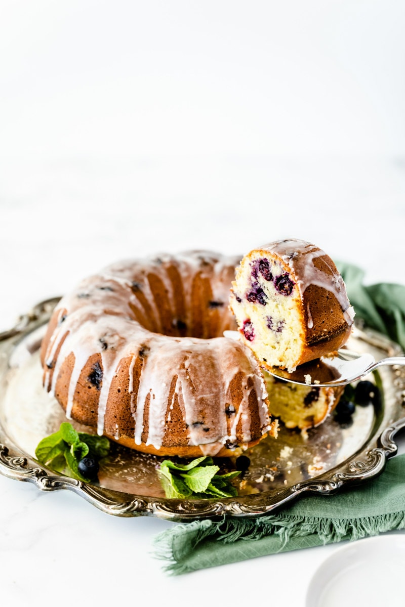 glazed blueberry lime pound cake on a silver platter. Showing a serving spatula lifting out a piece of the cake.