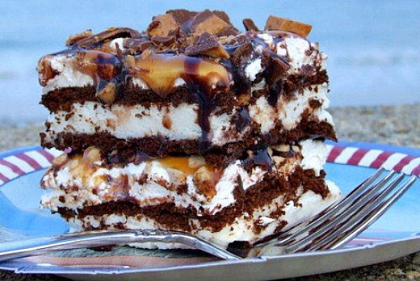 Frozen Caramel Toffee Ice Cream Sandwich Dessert recipe - from RecipeGirl.com #frozen #caramel #toffee #icecream #dessert #recipe #RecipeGirl slice of frozen caramel toffee ice cream sandwich dessert on a blue plate with red and white edge. Fork on the plate. Ocean in the background