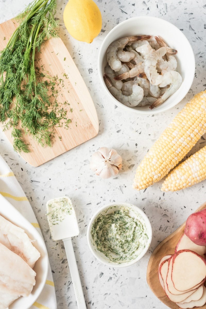 overhead shot of ingredients needed for new england seafood packets- herbs on a cutting board, raw shrimp in a white bowl, two corn on the cob, garlic bulb, white bowl of herb butter, sliced potatoes