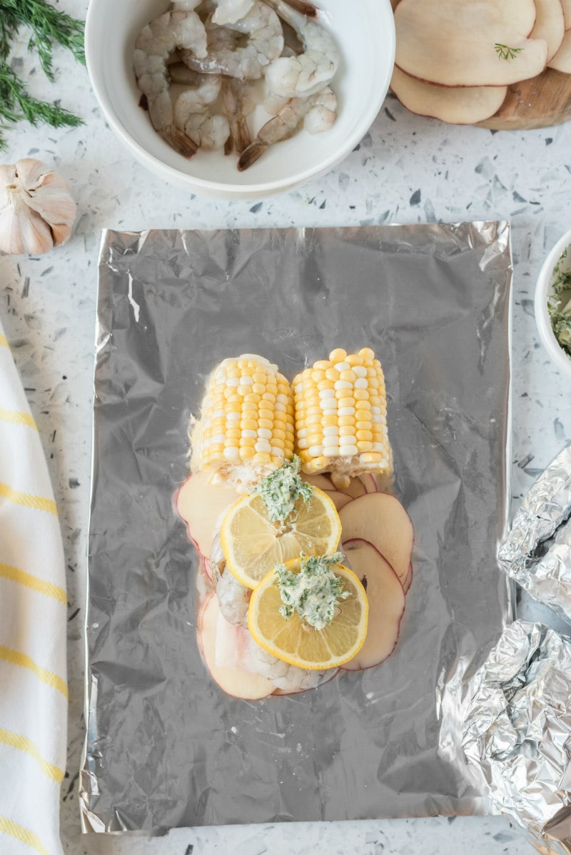 assembling seafood foil packets stacked with potato, fish, corn, shrimp and herb butter. white bowl of raw shrimp in the background