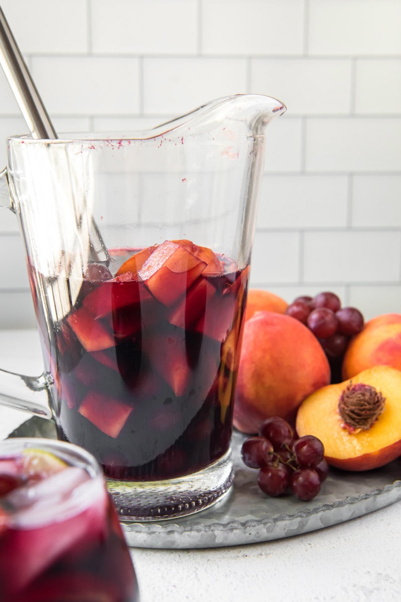 pitcher of red wine sangria with a stirring spoon inside. Grapes and peaches on the side