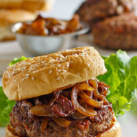 Bacon Burger with Balsamic Caramelized Onions on a white platter with a lettuce leaf. more buns, burgers and a bowl of caramelized onions in the background