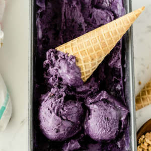 rectangular metal tub of blueberry cheesecake ice cream with a couple of scoops on top with an ice cream cone sitting in the tub