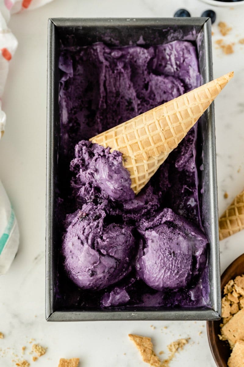rectangular metal tub of blueberry cheesecake ice cream with a couple of scoops on top with an ice cream cone sitting in the tub