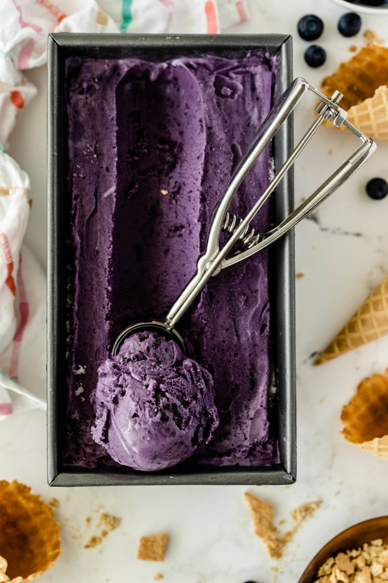 rectangular metal tub of blueberry cheesecake ice cream with an ice cream scoop that has scooped up some of the ice cream sitting on top of the tub