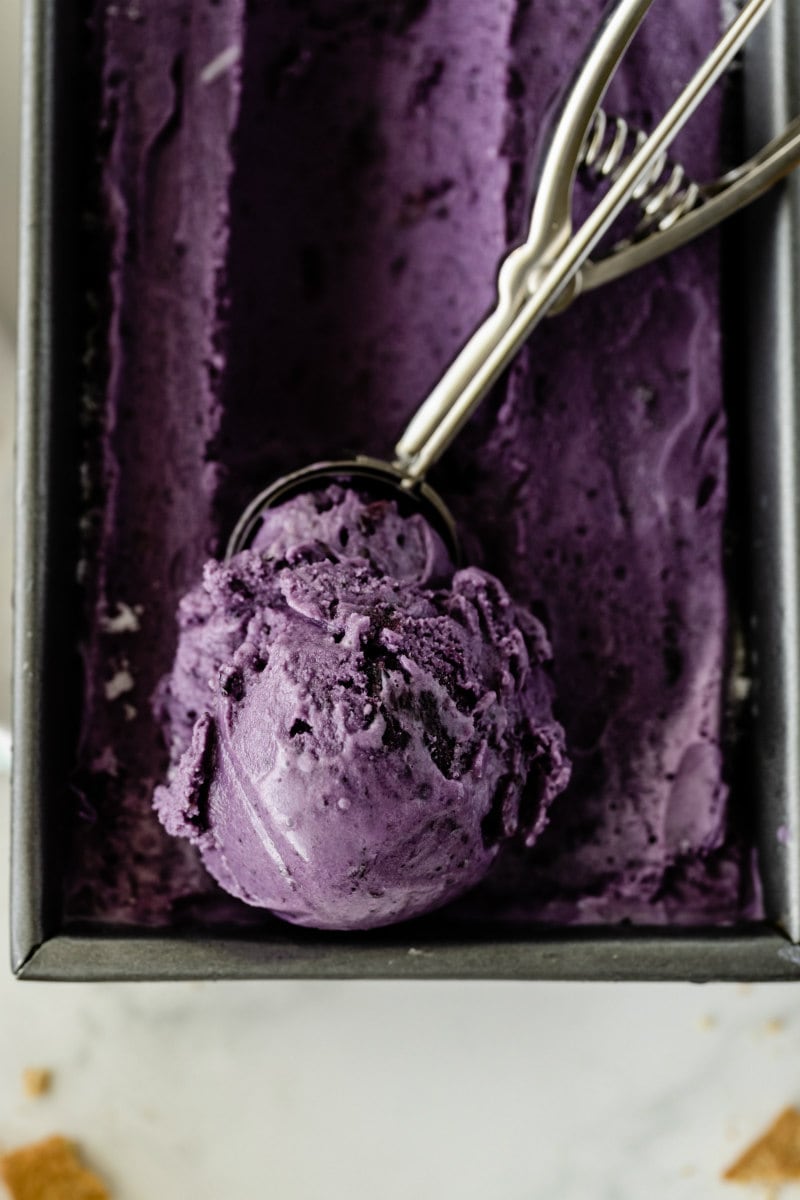 rectangular metal tub of blueberry cheesecake ice cream with an ice cream scoop that has scooped up some of the ice cream sitting on top of the tub