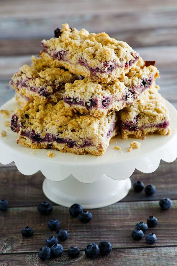 Lemon Blueberry Streusel Bars stacked on a white cake platter with fresh blueberries scattered underneath. Wood surface in the background.