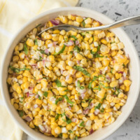 overhead shot of fresh corn salad in a white bowl with a serving spoon. light yellow napkin displayed on the side