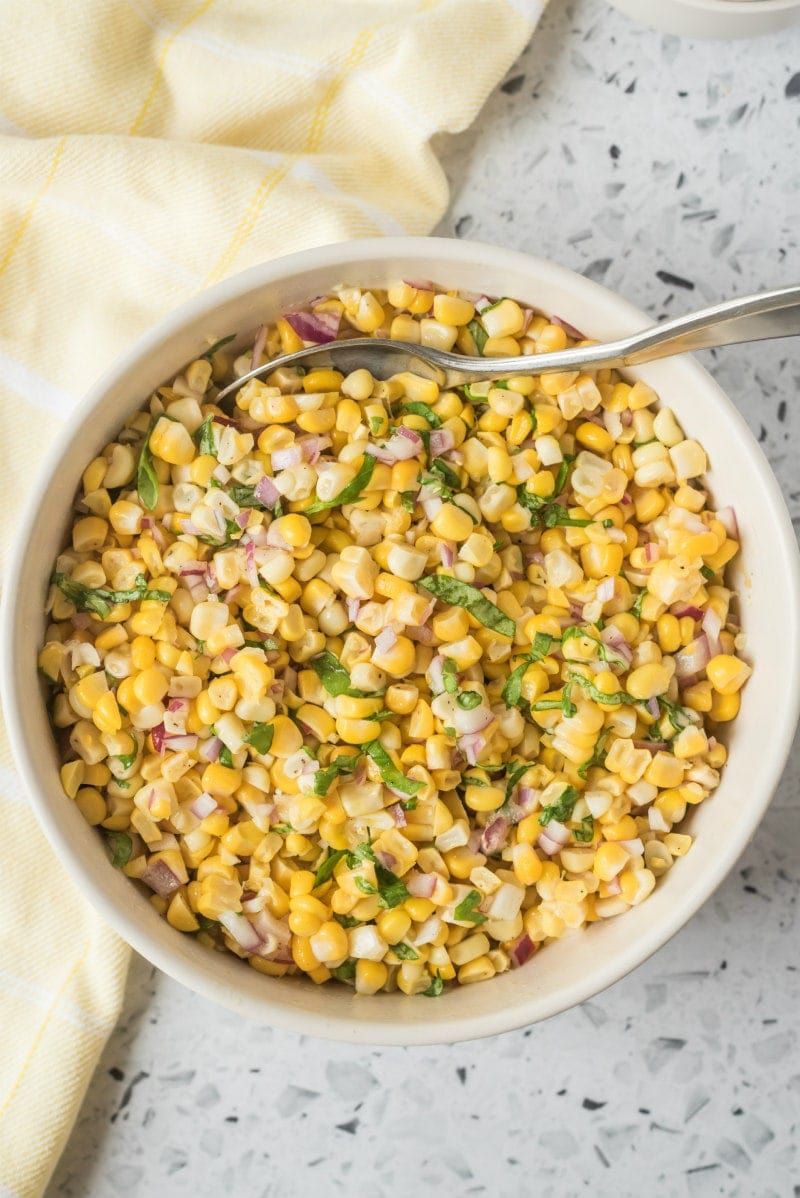 overhead shot of fresh corn salad in a white bowl with a serving spoon. light yellow napkin displayed on the side